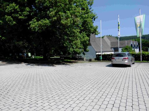 Ein Parkplatz mit gepflastertem Boden und einem Auto. Im Hintergrund sind Bäume und ein Gebäude mit Fahnen zu sehen.