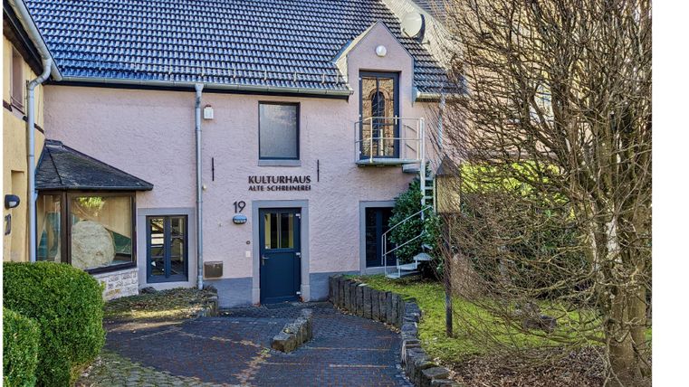 Eingang des Kulturhauses Alte Schreinerei mit gepflastertem Weg, Treppe und kahlem Baum im Vordergrund.