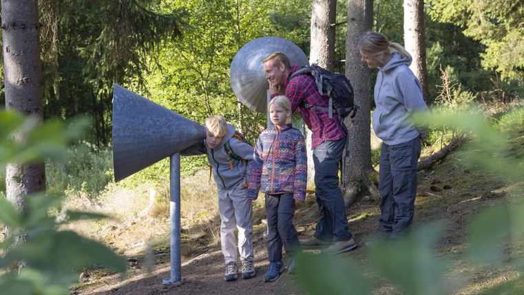 Eine Familie entdeckt ein akustisches Spielgerät im Wald. Die Kinder hören aufmerksam durch die großen, trichterförmigen Hörgeräte.