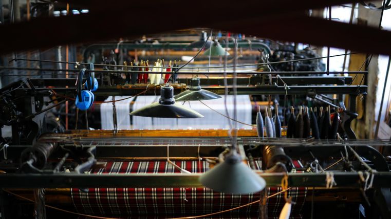 An old weaving machine with several spools and lamps can be seen. The woven fabric has a checkered pattern, and the surroundings are designed industrially.
