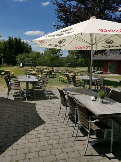 Een buitenruimte met tafels en stoelen, omgeven door een groene landschap. Parasols bieden schaduw op een zonnige dag.