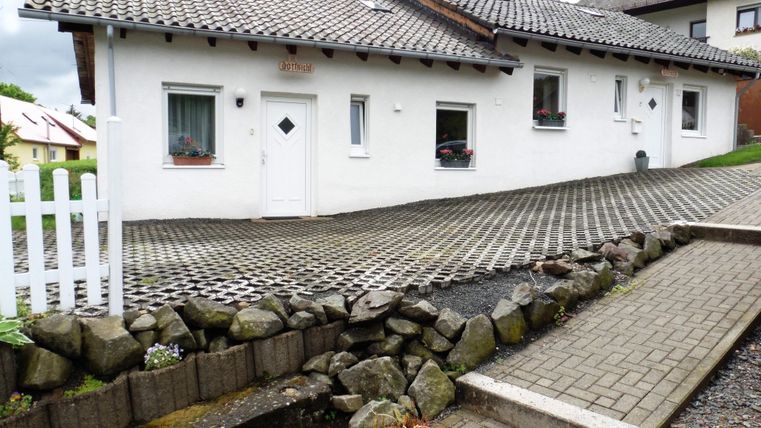 Ein modernes, weißes Haus mit einem steilen Dach und mehreren Fenstern. Die Auffahrt ist gepflastert und von einer kleinen Steinmauer umgeben.