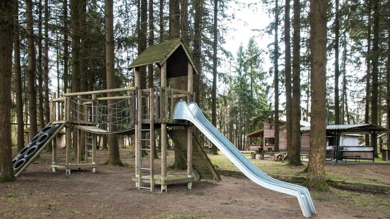 Een speeltuin in het bos met een houten speeltoestel en een glijbaan. Omgeven door hoge bomen is er ook een gebied met picknicktafels.