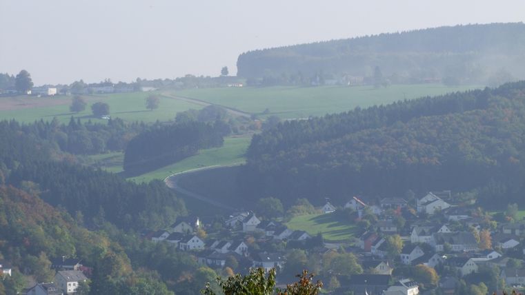 Eine malerische Landschaft mit sanften Hügeln und einem kleinen Dorf im Vordergrund. Die Umgebung ist grün und bewaldet, ideal für Naturliebhaber.