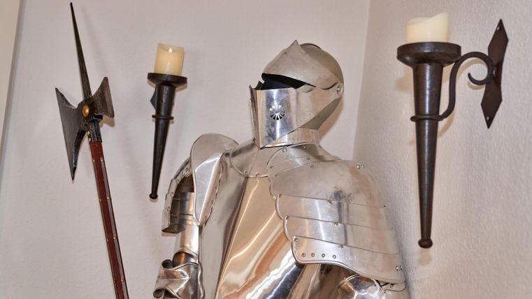 A silver knight's armor stand inside a room. Torches and a spearhead are mounted on the wall.