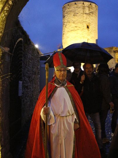 Een persoon in traditionele kleding met een rode cape staat onder een paraplu. Op de achtergrond is een verlichte toren te zien die een feestelijke sfeer creëert.