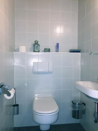 Ein modernes Badezimmer mit Toilette und Waschbecken. Die Wände sind hell gefliest und es sind einige dekorative Gegenstände auf dem Regal.