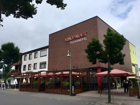 Ein modernes Gebäude mit dem Schild „Kino Palast Vulkaneifel“. Im Vordergrund befinden sich Terrassen mit Sonnenschirmen und Pflanzen.