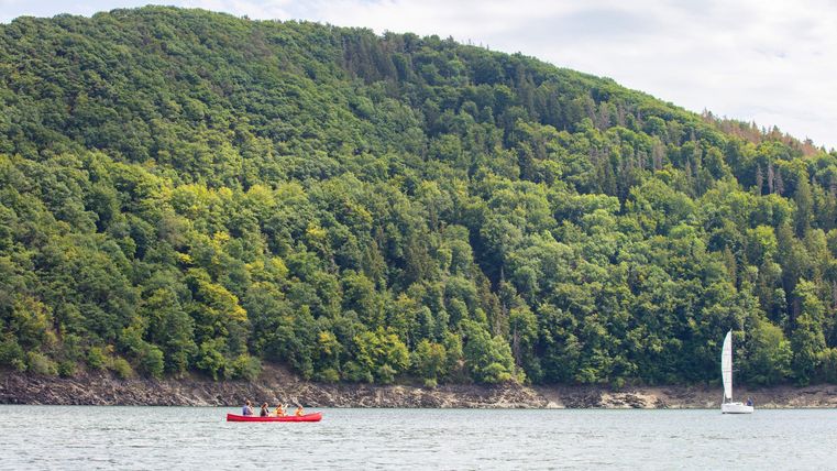 Een rustige meer omringd door groene heuvels. Een rode kajak en een zeilboot zijn zichtbaar op het water.