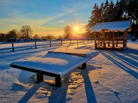 Eine verschneite Landschaft im Sonnenuntergang, mit einer Bank und einem Holzpavillon. Die langen Schatten der Bäume verleihen der Szene eine ruhige Atmosphäre.