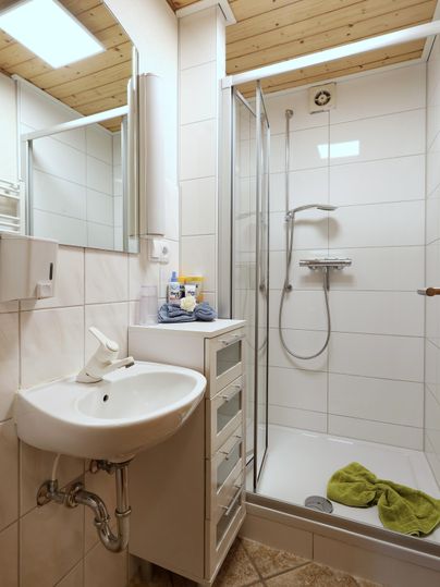 A modern bathroom with a shower and a sink. The walls are tiled in light colors, and a towel lies on the floor.