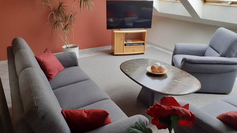 Een gezellige woonkamer met grijze banken en een ronde tafel. Grote ramen laten veel licht binnenstromen.