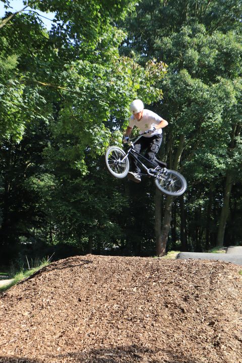 A BMX rider jumps over a ramp on a skate park hill. In the background, trees and a green environment can be seen.