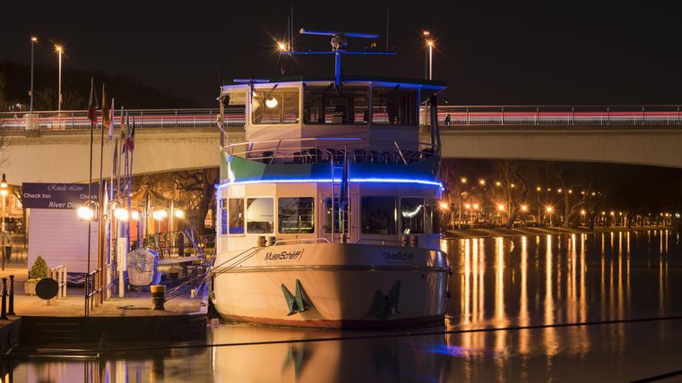 A illuminated boat lies by the shore at night. The lights reflect on the water, creating a calm atmosphere.