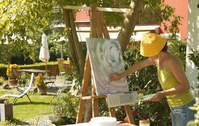 Eine Person malt an einer Leinwand im Freien. Im Hintergrund sind Gartenmöbel und Pflanzen zu sehen.