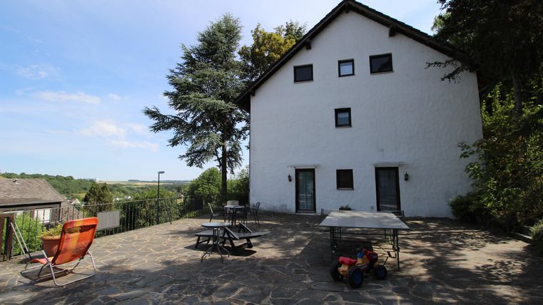 Ein weißes Haus mit einem gepflasterten Außenbereich und Stühlen. Im Hintergrund sind Bäume und eine grüne Landschaft zu sehen.