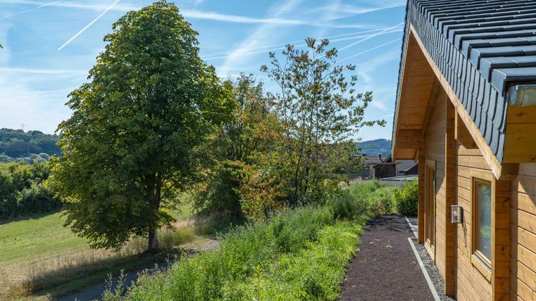 Ein schönes Haus mit Holzfassade und einem gepflegten Garten. Im Hintergrund sind grüne Wiesen und ein klarer blauer Himmel zu sehen.