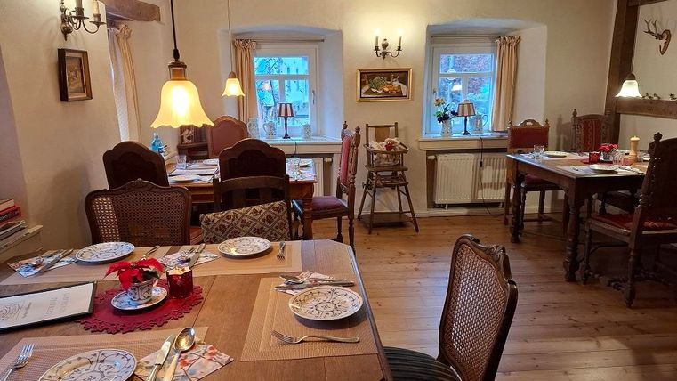 A cozy dining area with wooden tables and chairs. The tables are lovingly set with plates and cutlery, and there are windows with curtains that create an inviting atmosphere.