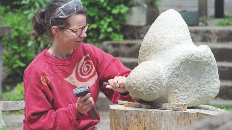 An artist is working on a stone block with a hammer and chisel. In the background, there are green plants and a relaxed environment.
