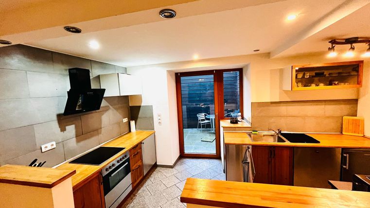 A modern kitchen with wood and stone surfaces. The large windows offer a view of an outdoor area with seating.