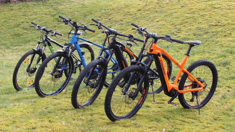 Five mountain bikes are on a green meadow. The bicycles are in different colors, including black, blue, green, and orange.