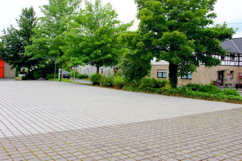 An empty square with a paved floor and some trees. Buildings can be seen in the background.
