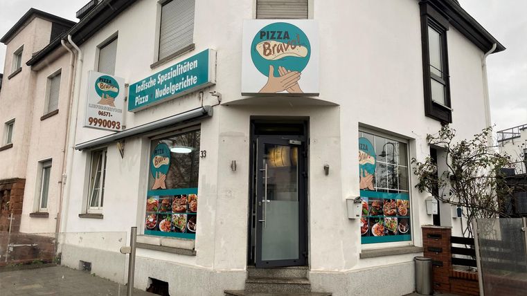 Ein Restaurant mit einer hellen Fassade und großen Fenstern. Die Schilder zeigen verschiedene Speisen und das Ambiente wirkt einladend.