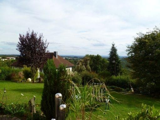 Eine ruhige Gartenlandschaft mit einem Haus und üppigem Grün. Im Hintergrund sind Wälder und ein blauer Himmel zu sehen.
