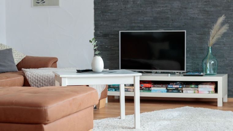 Ein gemütlicher Wohnbereich mit einem braunen Sofa und einem weißen Couchtisch. Im Hintergrund steht ein Fernseher und eine Regale mit Büchern.