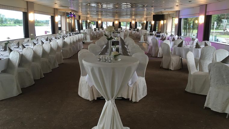 An elegant event space with white tables and chairs. Large windows offer a view of the water.