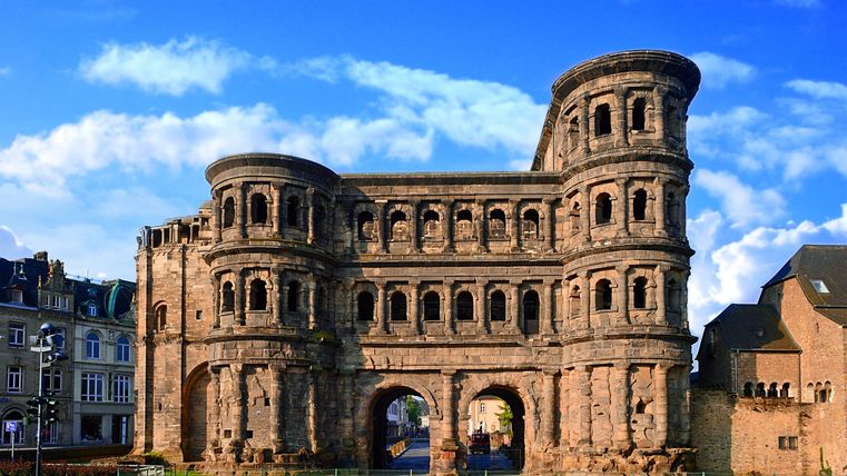 Ein historisches Bauwerk mit einer beeindruckenden Fassade und mehreren Torbögen. Der Himmel ist klar und blau mit einigen Wolken.