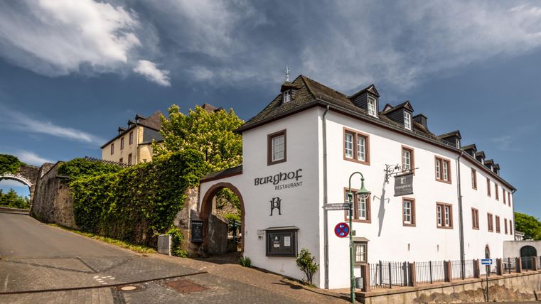 Een historisch gebouw met een lichte façade en een mooie ingang. Op de achtergrond zijn groene bomen en een blauwe lucht te zien.
