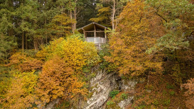 Eine Aussichtshütte auf einem Felsen, umgeben von buntem Herbstlaub. Die Farben der Blätter variieren zwischen Gelb, Orange und Grün.