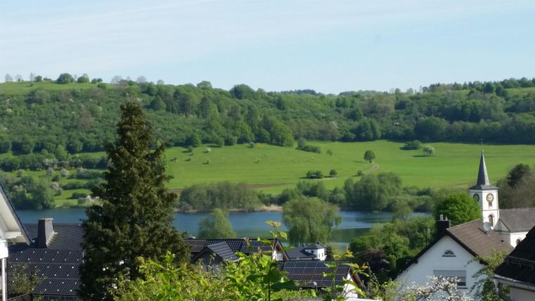 Eine malerische Landschaft mit grünen Wiesen und einem ruhigen See. Im Hintergrund stehen sanfte Hügel und ein Kirchturm ist sichtbar.
