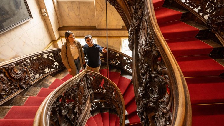 Een elegante trap met een rood gekleurd tapijt en een kunstzinnig houten leuningen. Twee personen staan en kijken omhoog.