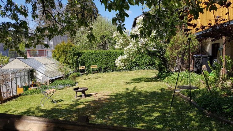 Een groene tuin met een grasveld en bloeiende bomen. Op de achtergrond bevinden zich een tuinhuis en twee banken.