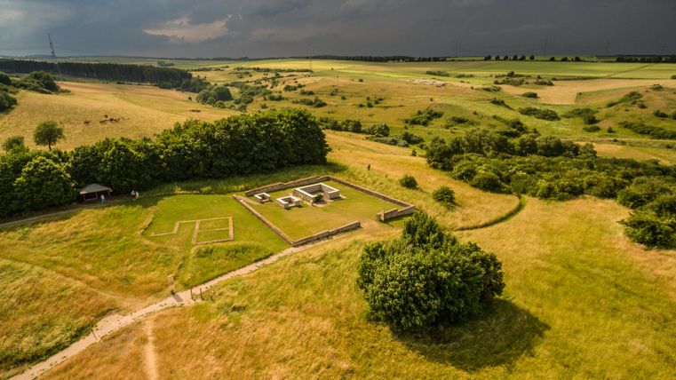 Eine weite Landschaft mit grünen Feldern und bewaldeten Bereichen. In der Mitte sind Überreste einer historischen Stätte sichtbar.