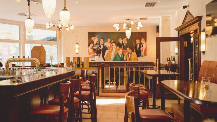 Een elegante bar met houten stoelen en een groot schilderij aan de muur. De ruimte is helder verlicht en straalt een uitnodigende sfeer uit.