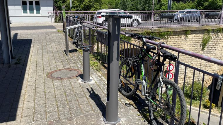 Een fietsenrek op een trottoir, met meerdere fietsen die veilig zijn afgesloten. Op de achtergrond is een groen landschap en een paar auto's te zien.