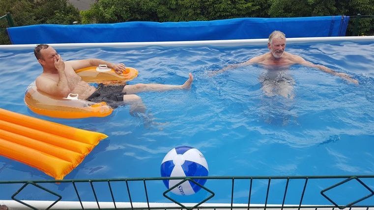Zwei Männer entspannen in einem Pool. Einer sitzt auf einer Luftmatratze, während der andere im Wasser plantscht.