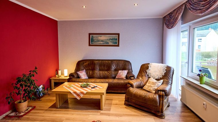 Ein gemütlicher Wohnraum mit braunen Ledermöbeln und einem Holzkaffee-Tisch. Eine rote Wand und ein Fenster sorgen für eine einladende Atmosphäre.