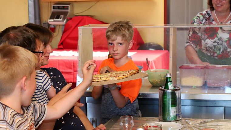 A group of children is sitting at a table and looking at a boy holding a pizza on a plate. In the background, there is a woman who is working in the kitchen.