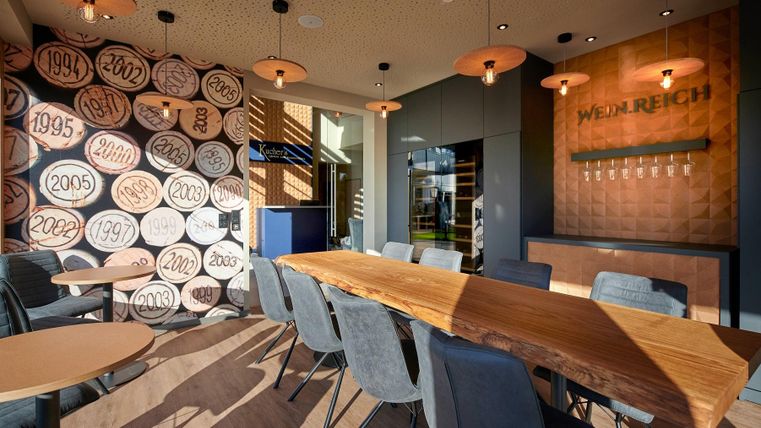 Ein modernes Restaurant mit einem großen Holztisch und stilvollen Stühlen. Die Wände sind mit dekorativen Fässern und einer beeindruckenden Anzeigetafel gestaltet.