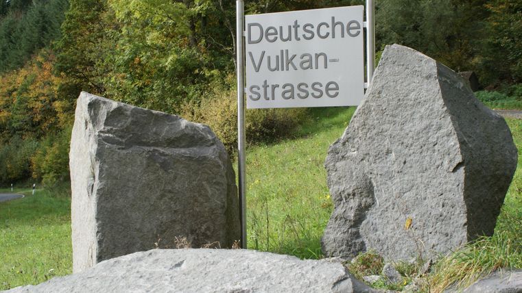 Ein Schild mit der Aufschrift "Deutsche Vulkanstraße" steht zwischen großen Steinen. Im Hintergrund sind Bäume in herbstlichen Farben zu sehen.
