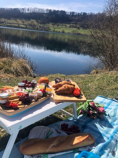 A beautifully set picnic table by the shore of a tranquil lake. On the table, various foods, bread, and flowers can be seen.