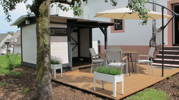 Eine gemütliche Terrasse mit einem Tisch und Stühlen unter einem Sonnenschirm. Im Hintergrund steht ein kleiner Schuppen neben einem Haus.