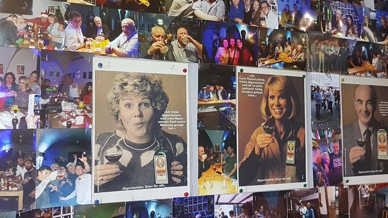 Een muur vol foto’s van mensen op feesten en evenementen. Sommige foto’s tonen personen met drankjes in de handen, terwijl andere de sfeer van het feest vastleggen.