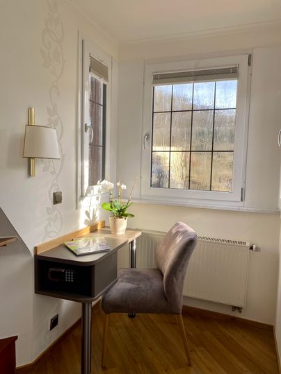 Ein gemütlicher Arbeitsplatz mit einem Schreibtisch und einem Stuhl. Am Fenster steht eine blühende Pflanze und bietet einen Blick nach draußen.