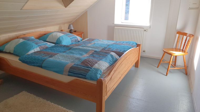 Een heldere slaapkamer met een houten bed en kleurrijke dekbedden. Naast het bed staat een houten stoel en een raam laat daglicht binnen.