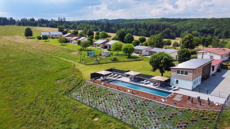 Een rustige landschap met een modern huis en een zwembad. Op de achtergrond zijn verschillende kleinere gebouwen en veel groen te zien.